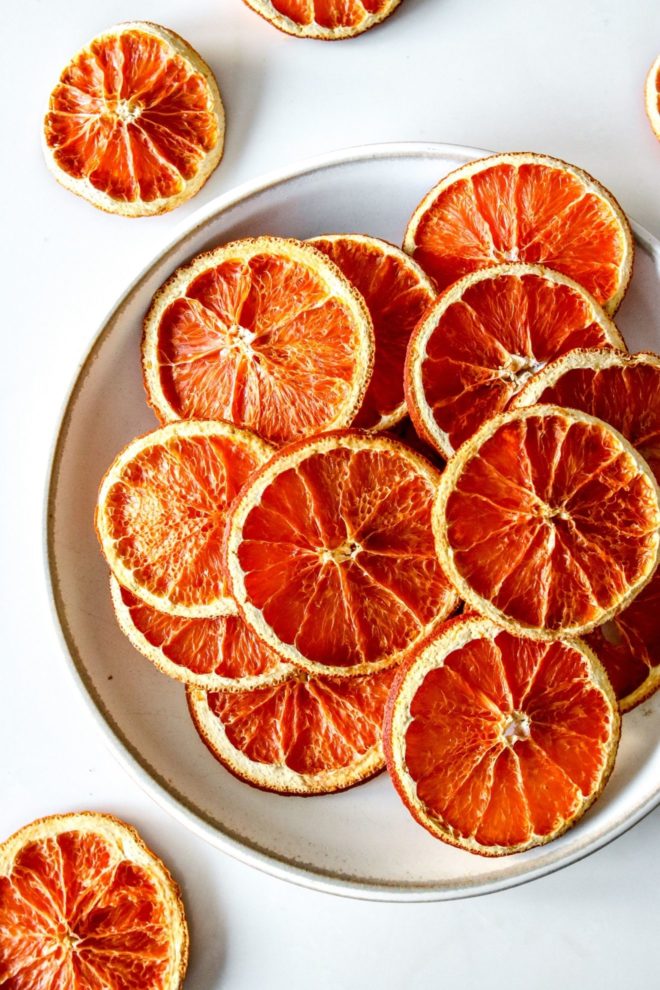 This is an overhead image of a white plate with dried orange slices on it. The plate sits on a white counter with more orange slices on the counter around the plate.