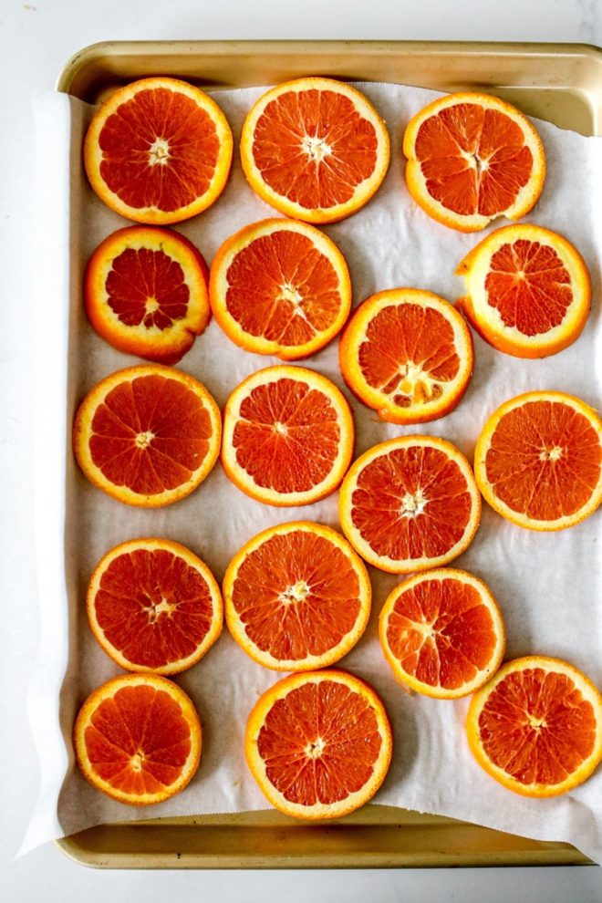 This is an overhead image of a gold baking sheet with white parchment paper and orange slices laid out in an even layer. The baking sheet sits on a white counter.
