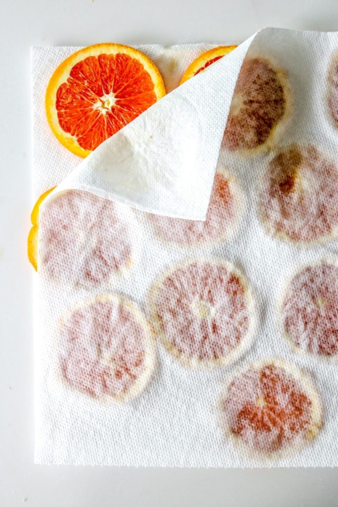 This is an overhead image of oranges slices in between two sheets of a paper towel. The top paper towel is peeled back a little bit to reveal part of an orange slice. The paper towels sit on a white counter.