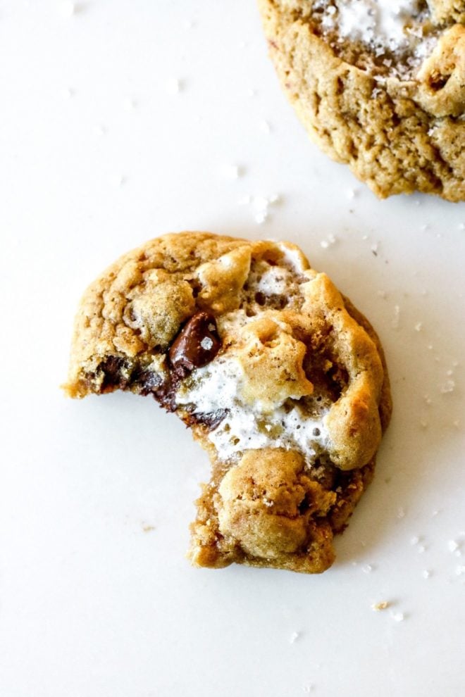 This is an overhead image of a cookie with chocolate chips and marshmallows. The cookie has a bite taken out of it and sits on a white counter with another cookie in the top right and bottom right corner of the image.