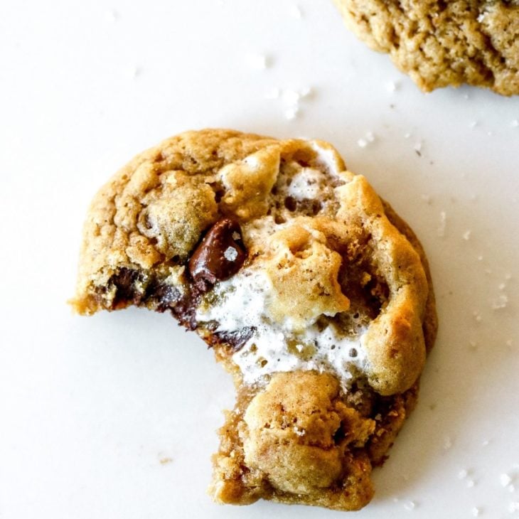 This is an overhead image of a cookie with chocolate chips and marshmallows. The cookie has a bite taken out of it and sits on a white counter with another cookie in the top right and bottom right corner of the image.