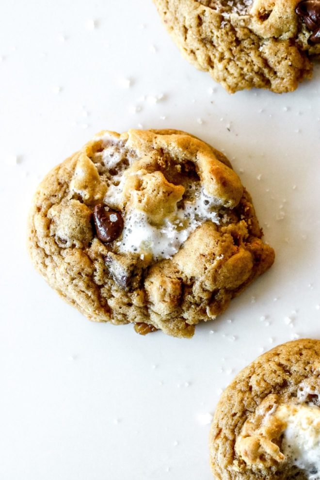 This is an overhead image of a cookie with chocolate chips and marshmallows. The cookie sits on a white counter with another cookie in the top right and bottom right corner of the image.
