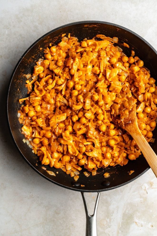 This is an overhead image of a black skillet with a chickpea and jackfruit mixture with orange buffalo sauce. The skillet has a wooden spatula in it and leaning against the right side of the skillet. The skillet sits on a light white surface.