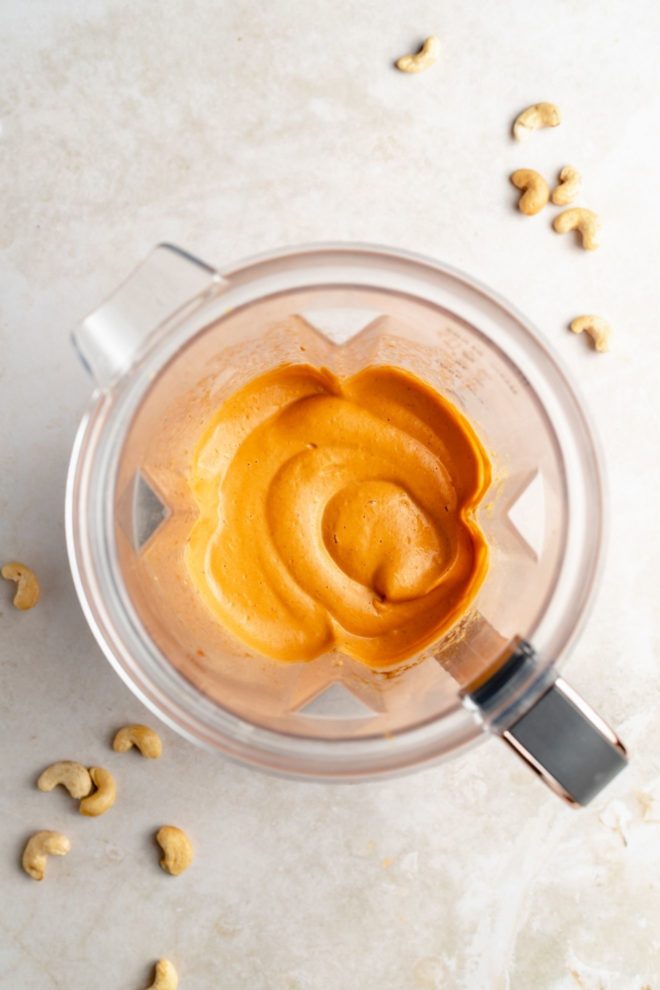 This is an overhead image of a blender with blended queso in it. The blender sits on a light white/beige surface with some raw cashews sprinkled around it.