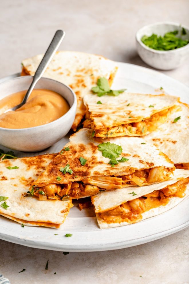 This is a side view looking onto a white plate. On the plate is cut triangles of a quesadilla with chickpeas and jackfruit. A small bowl of queso is on the plate with a spoon sticking out of it. The plate sits on a light tan surface with a small bowl of cilantro blurred in the background.