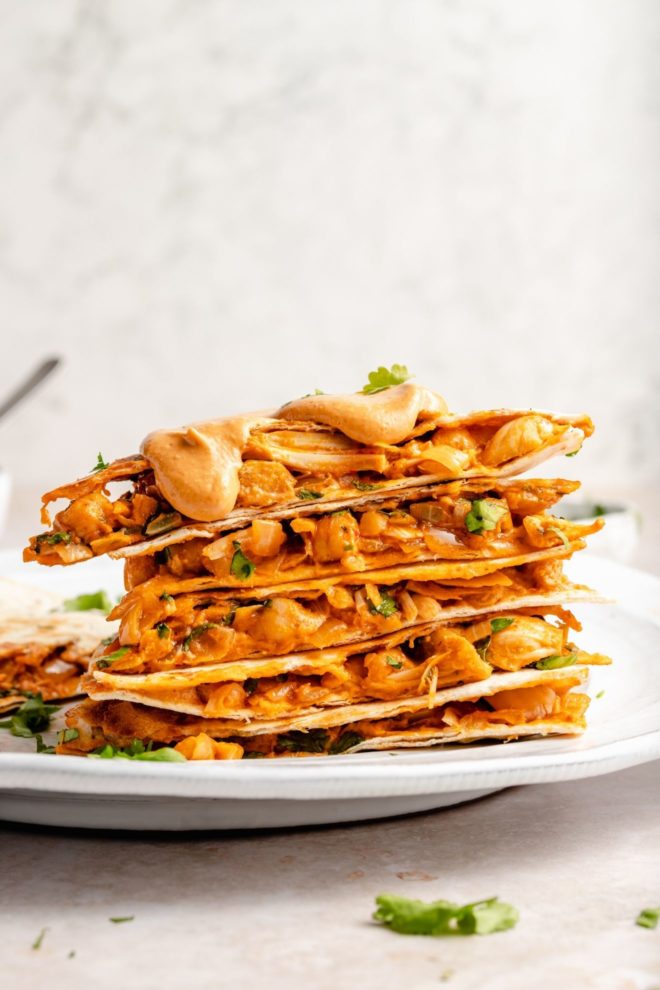 This is a side view of a stack of quesadilla sliced. The quesadilla has chickpeas, jackfruit, and little pieces of cilantro. The stack is topped with a queso. The stack of quesadilla pieces are on a white plate on a white surface with a white background.