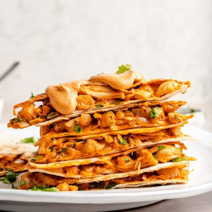 This is a side view of a stack of quesadilla sliced. The quesadilla has chickpeas, jackfruit, and little pieces of cilantro. The stack is topped with a queso. The stack of quesadilla pieces are on a white plate on a white surface with a white background.