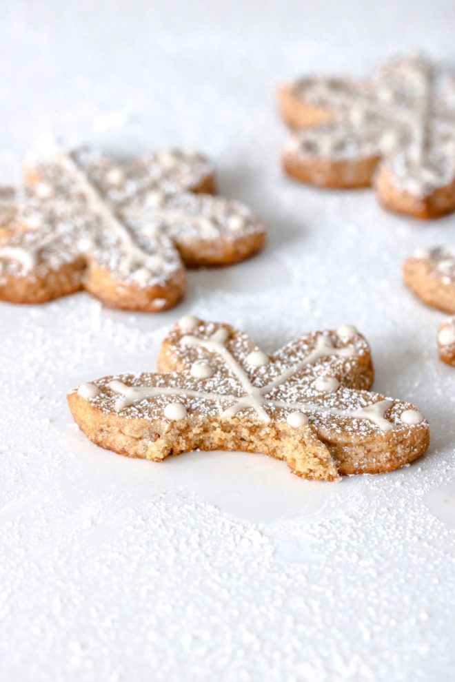 This is a side view of a shortbread snowflake cutout cookie with a bite taken out of it. The cookies sit on a white surface. The cookies are decorated with vanilla royal icing and sprinkled with powdered sugar.
