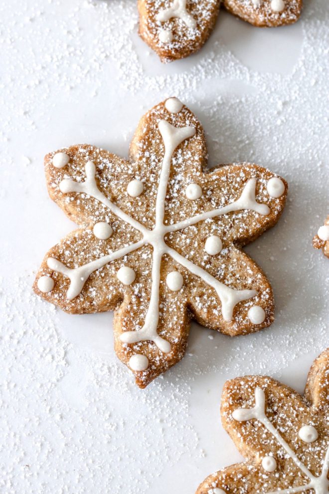 This is an overhead image of a shortbread snowflake cutout cookie. The cookie is on a white surface with another cookie to the top of the image and another cookie to the bottom right of the image. The cookie is decorated with white royal icing and sprinkled with powdered sugar.