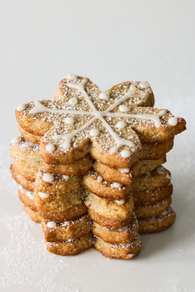 This is a stack of shortbread snowflake cutout cookies. The stack sits on a white surface. The cookies are decorated with a white royal icing and sprinkled with powdered sugar.