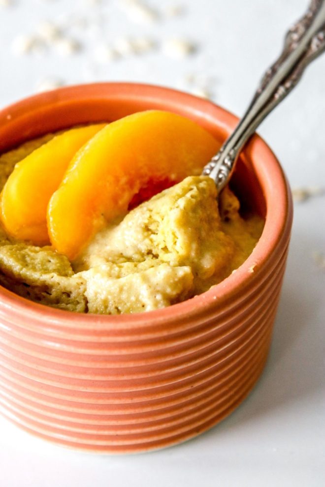 This is a side view of peach baked oatmeal in a coral bowl. An antique spoon is dipping into the oats and peach slices are on top. The bowl sits on a white counter with oats scattered around the bowl.