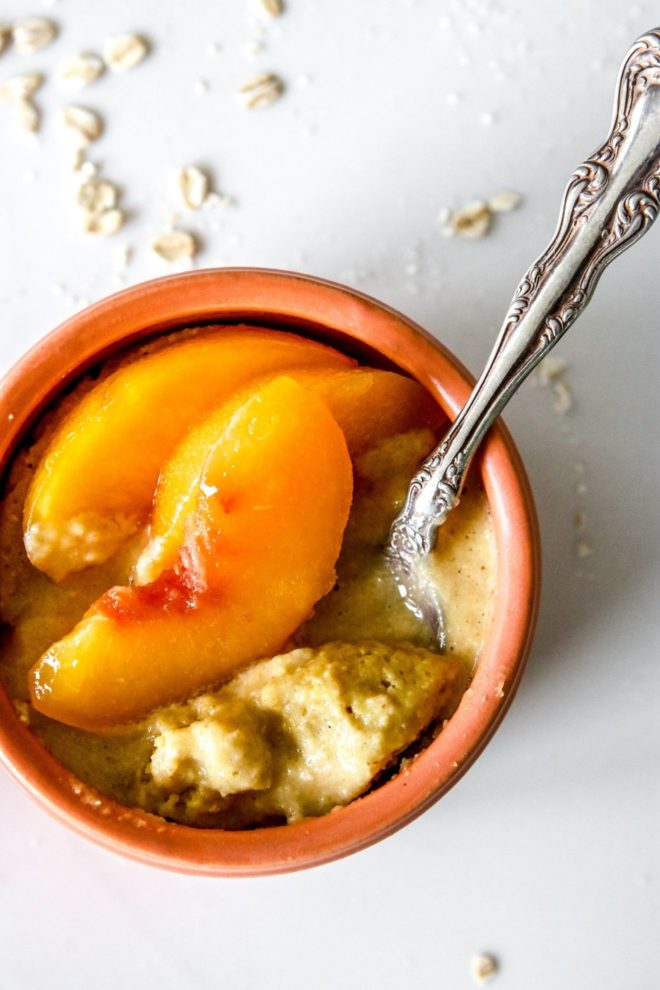 This is an overhead image of peach baked oatmeal in a coral bowl. An antique spoon is dipping into the oats and peach slices are on top. The bowl sits on a white counter with oats scattered around the bowl.