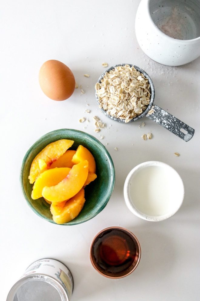 This is an overhead image of ingredients to make peach baked oatmeal. Salt, an egg, oats, peach slices, milk, agave nectar, baking powder are on a white counter.