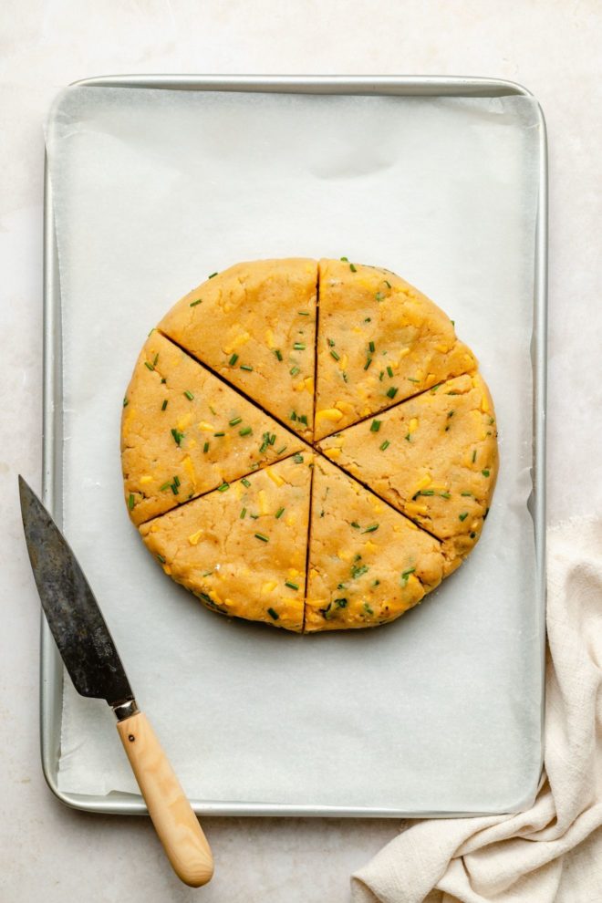 This is an overhead image of a baking sheet with white parchment paper. On the parchment paper is a disc of raw scones dough with flecks of cheese and chives. The dough is cut into 6 triangles and a knife with a wooden handle is leaning against the side of the baking sheet. A beige tea towel is in the bottom right corner of the image.