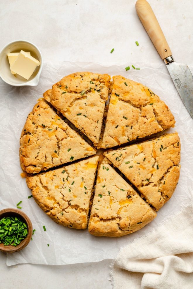 This is an overhead image of cooked scones with bites of cheese and chives. The scones are cut into triangles and on top of a piece of white parchment paper. A knife is on the top right corner of the image. A small bowl of butter is to the top left of the scones. A small bowl of chives to the bottom left of the scones and a beige tea towel to the bottom right of the image.