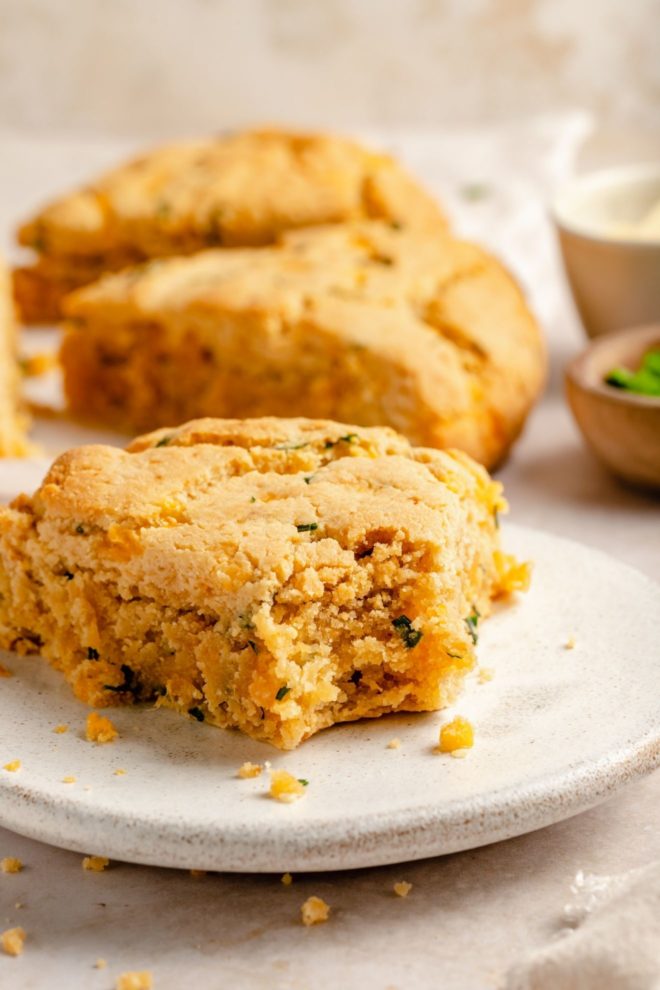 This is a side view of a cheese and chive scone with a bite taken out of it. The scone sits on a white plate which is sitting on a beige surface. More scones are blurred in the background.