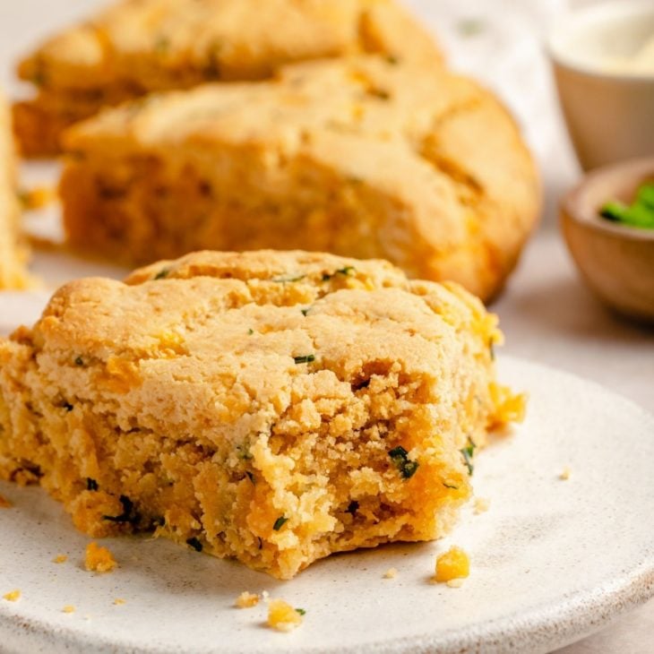 This is a side view of a cheese and chive scone with a bite taken out of it. The scone sits on a white plate which is sitting on a beige surface. More scones are blurred in the background.