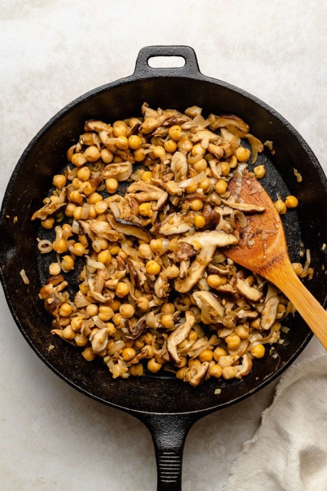 This is an overhead image of chickpeas, mushrooms, and shallots in a cast iron skillet. The skillet is on a white surface with a wood spatula. A tan tea towel is in the bottom right corner of the image.