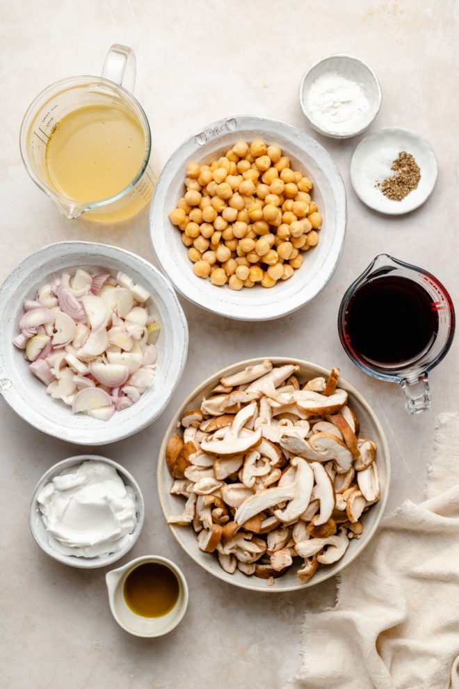 This is an overhead image of ingredients on a white surface. The ingredients are broth, chickpeas, salt, pepper, arrowroot flour, marsala wine, chopped shallots, sliced mushrooms, coconut cream and oil.