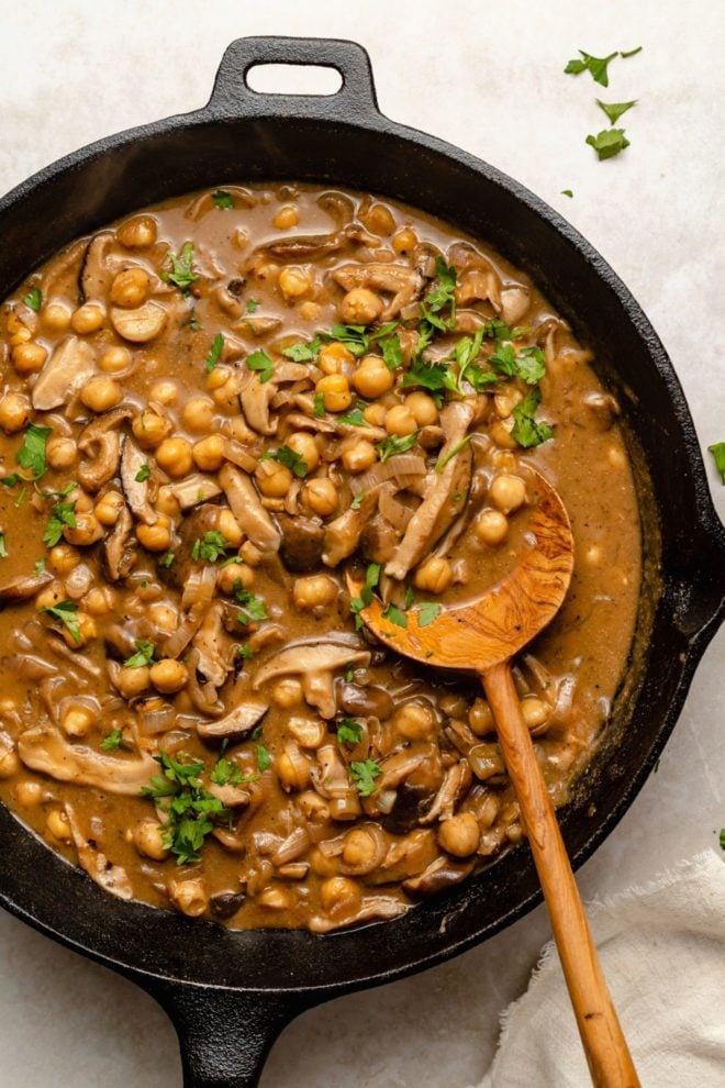 This is an overhead view of a cast iron skillet with chickpeas and mushrooms in a marsala sauce. A wood spoon is scooping up some of the chickpeas and leaning against the skillet. The chickpeas are topped with parsley leaves.