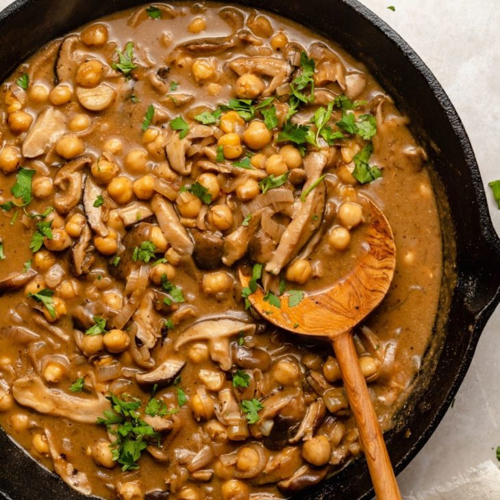 This is an overhead view of a cast iron skillet with chickpeas and mushrooms in a marsala sauce. A wood spoon is scooping up some of the chickpeas and leaning against the skillet. The chickpeas are topped with parsley leaves.