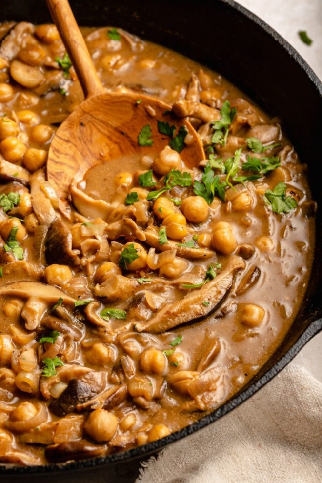This is a side view of a cast iron skillet with chickpeas and mushrooms in a marsala sauce. A wood spoon is scooping up some of the chickpeas and leaning against the skillet. The chickpeas are topped with parsley leaves.
