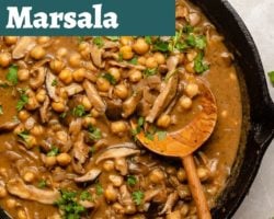 This is an overhead view of a cast iron skillet with chickpeas and mushrooms in a marsala sauce. A wood spoon is scooping up some of the chickpeas and leaning against the skillet. The chickpeas are topped with parsley leaves. Text overlay reads "vegan chickpea mushroom marsala."