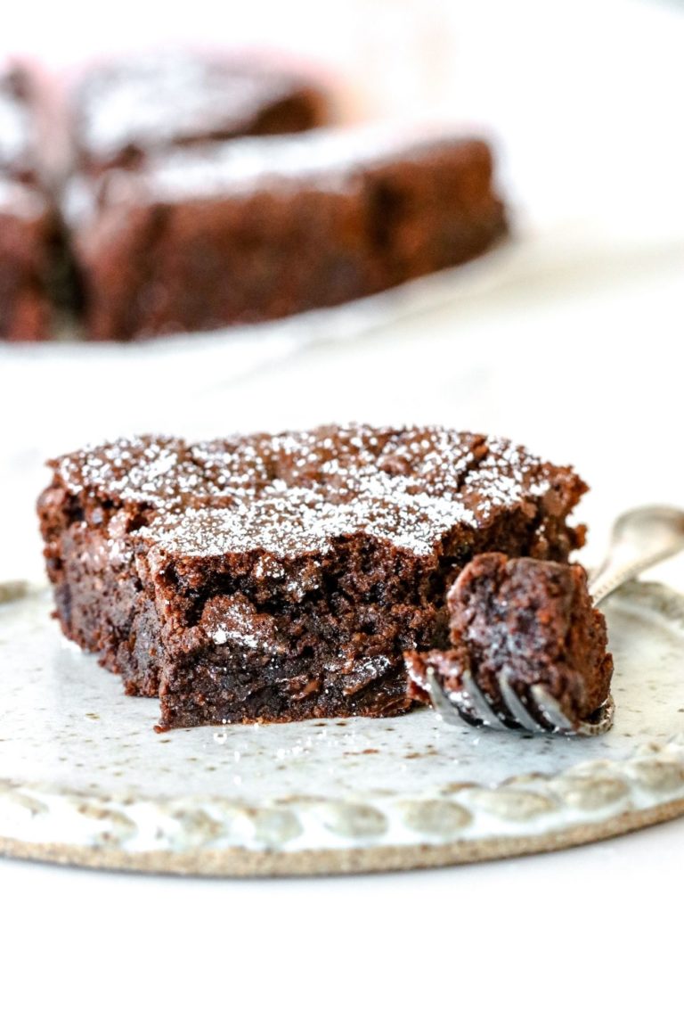 This is a side view of a chocolate cake slice on a plate. The plate is on a white counter and more cake is blurred in the background. A fork has a bite of the cake and is leaning on the side of the plate next to the slice of chocolate cake.
