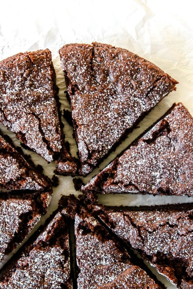 This is an overhead image of a chocolate cake cut into 8 slices. The image focuses on the top of the cake and is on a white piece of parchment paper. The slices of cake are pulled away from each other and sprinkled with powdered sugar.