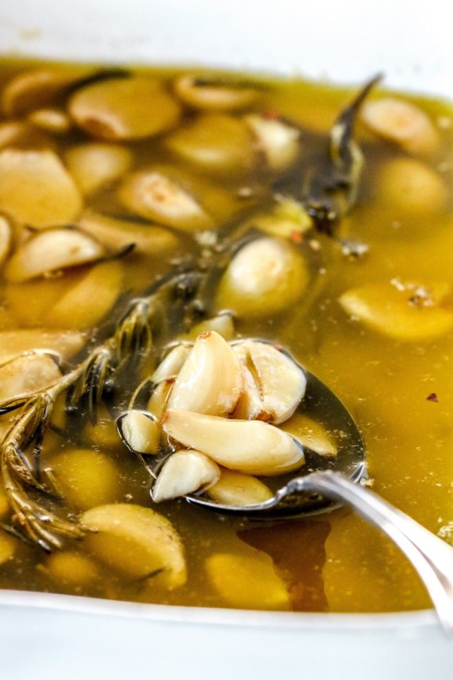 This is a side view of a dish of garlic confit. A spoon scooping up garlic cloves from oil. The garlic cloves and oil is in a white dish with more cloves and oil and a rosemary sprig.