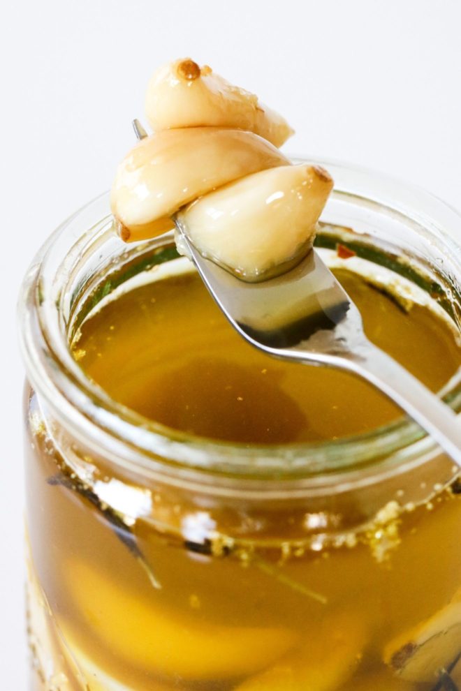This is a side view of a glass jar filled with garlic confit. A fork is piercing three garlic cloves and leaning against the side of the jar. The background of the image is white.