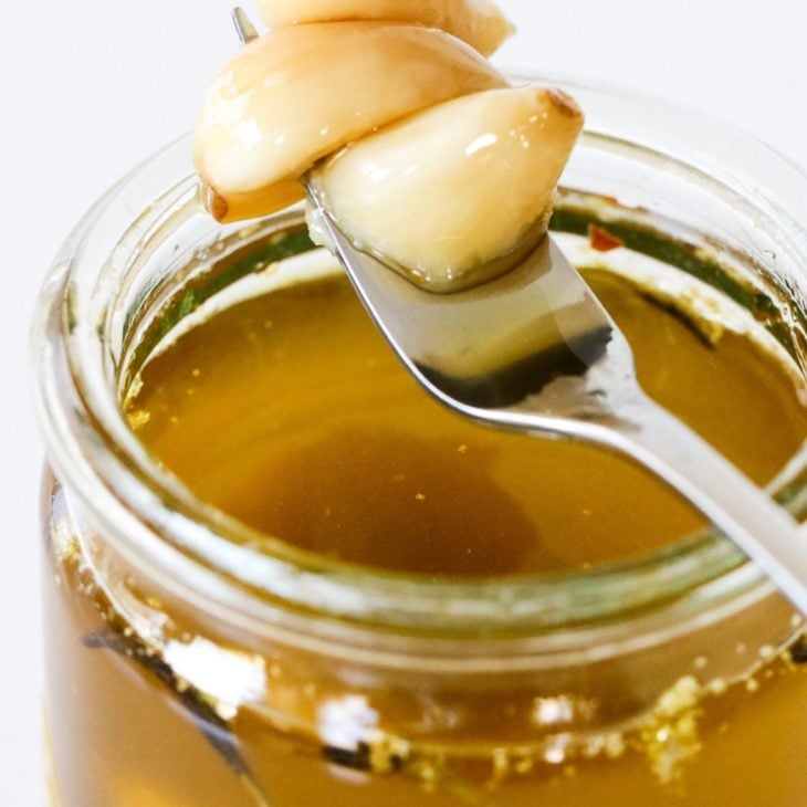 This is a side view of a glass jar filled with oil. A fork is piercing three garlic cloves and leaning against the side of the jar. The background of the image is white.