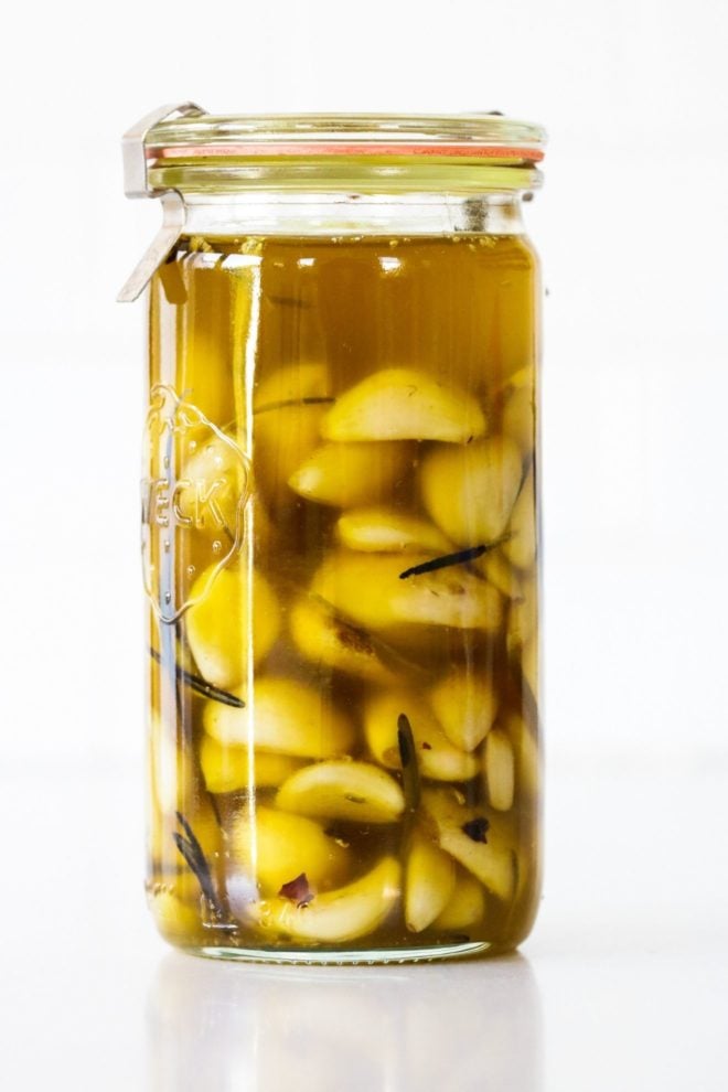 This is a side view of a tall glass jar filled with oil and garlic. The jar sits on a white counter with a white backhround.