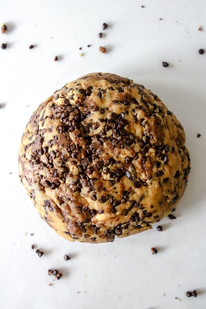 This is an overhead image of a ball of scone dough with mini chocolate chips. The scone disc sits on a white counter with chocolate chips around it.