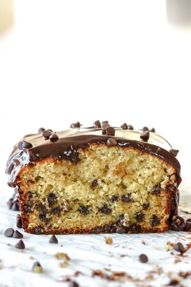 This is a side view of a chocolate chip loaf sliced into it. You can see the mini chocolate chips in the loaf. The loaf is topped with a chocolate ganache frosting with mini chocolate chips on top and around the loaf. The loaf sits on a white piece of parchment paper and a white background.