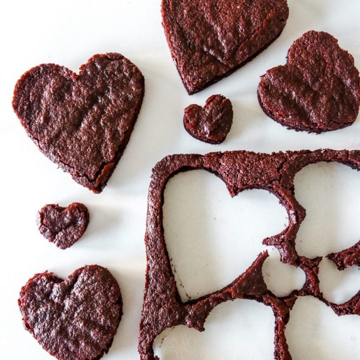 This is an overhead image of a sheet of chocolate cake with hearts cut out of it. The cake is to the bottom right of the image with heart shapes cut out of it. The chocolate heart cakes are around the sheet cake on a white counter.