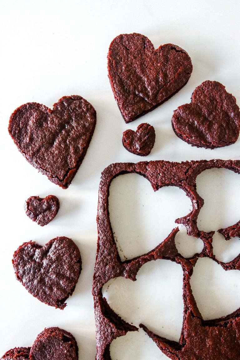 This is an overhead image of a sheet of chocolate cake with hearts cut out of it. The cake is to the bottom right of the image with heart shapes cut out of it. The chocolate heart cakes are around the sheet cake on a white counter.