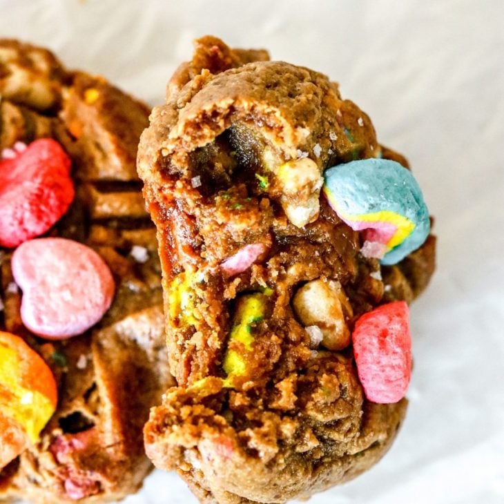 This is an overhead image of a peanut butter cookie with a bite taken out of it. The cookie is topped with colorful marshmallows and leaning against another cookie. The cookies sit on a white piece of parchment paper.