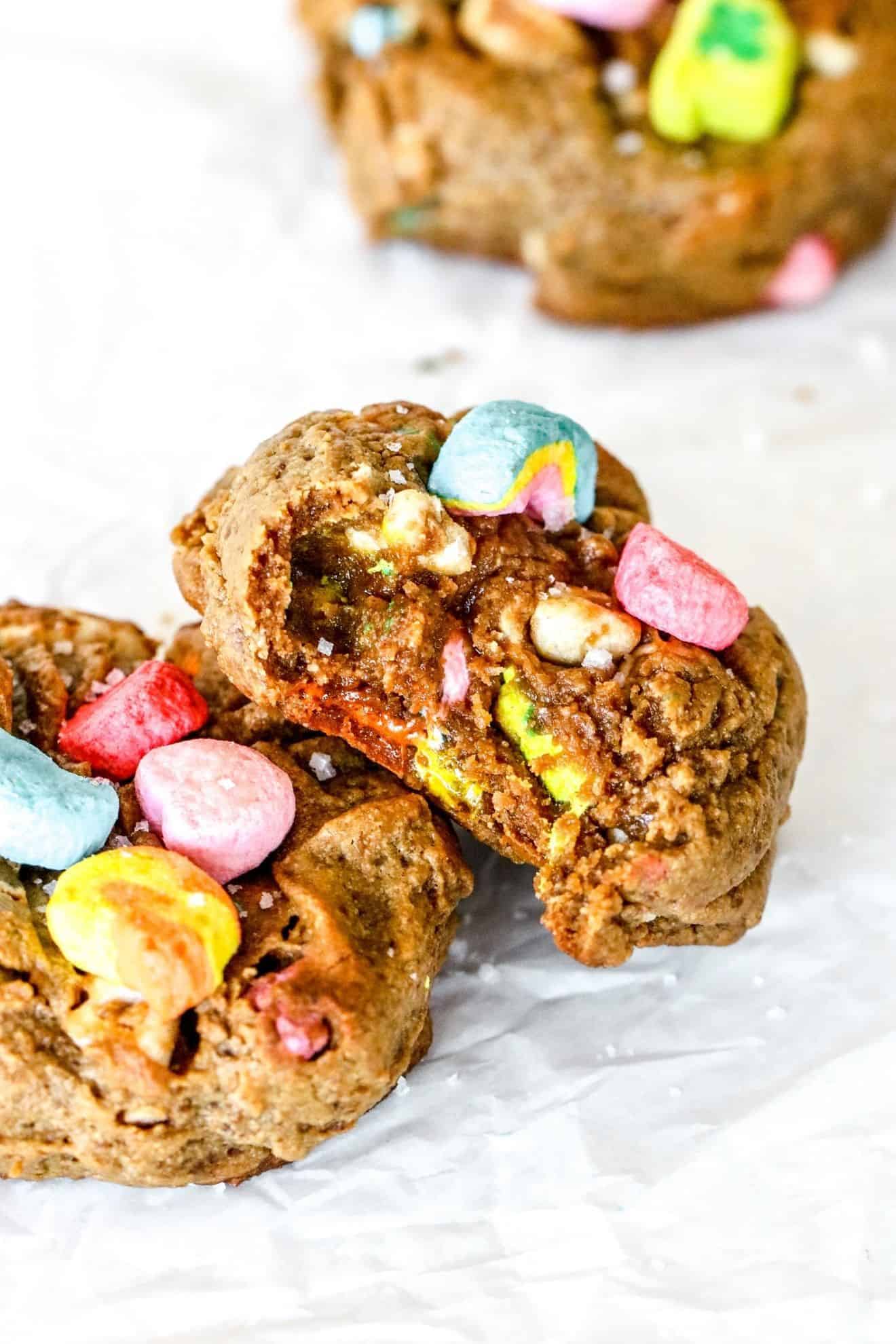 This is a side view image of a peanut butter cookie with a bite taken out of it. The cookie is topped with colorful marshmallows and leaning against another cookie. The cookies sit on a white piece of parchment paper. Another cookie is blurred in the right top corner of the image.