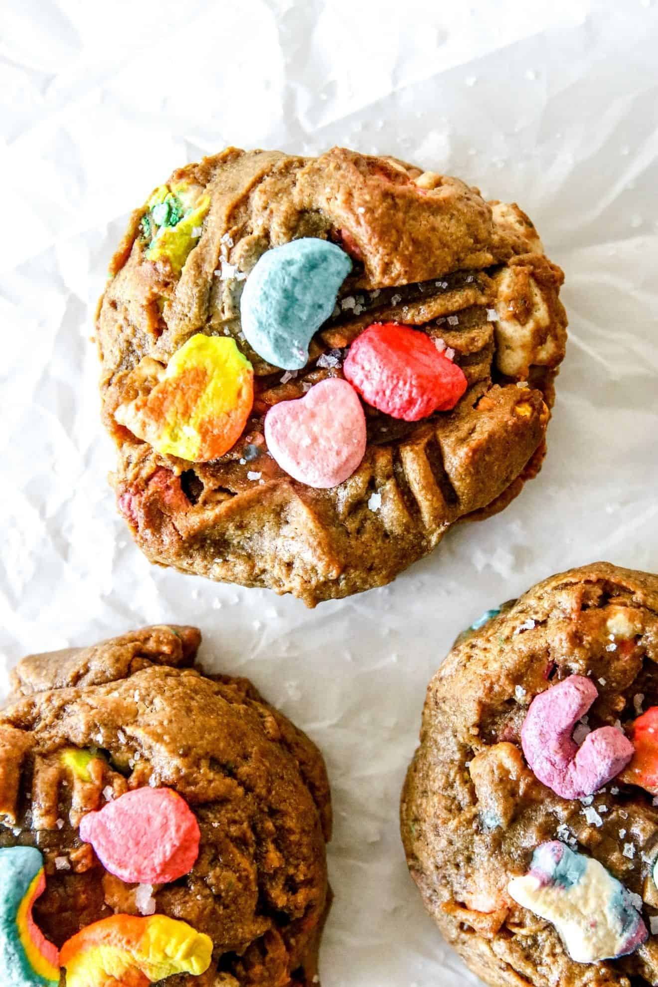 This is an overhead image of a peanut butter cookie topped with colorful lucky charms marshmallows. The cookie sits on a white piece of parchment paper with another cookie in the right top corner of the image.