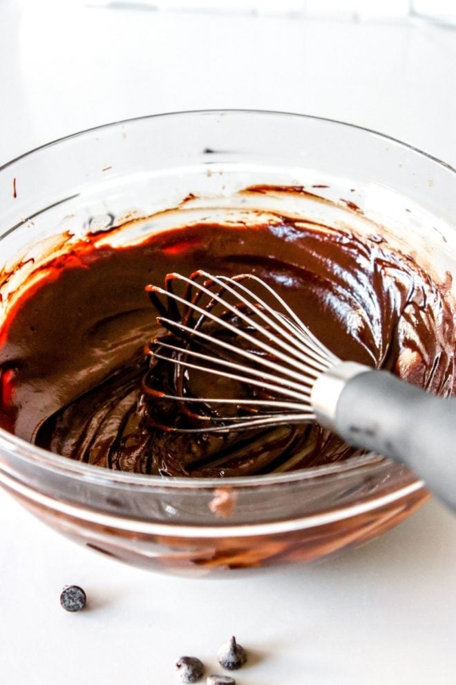 This is an angled view looking into a glass bowl with melted chocolate in it. A whisk is in the bowl leaning against the side. The bowl sits on a white counter with some chocolate chips in front of it.