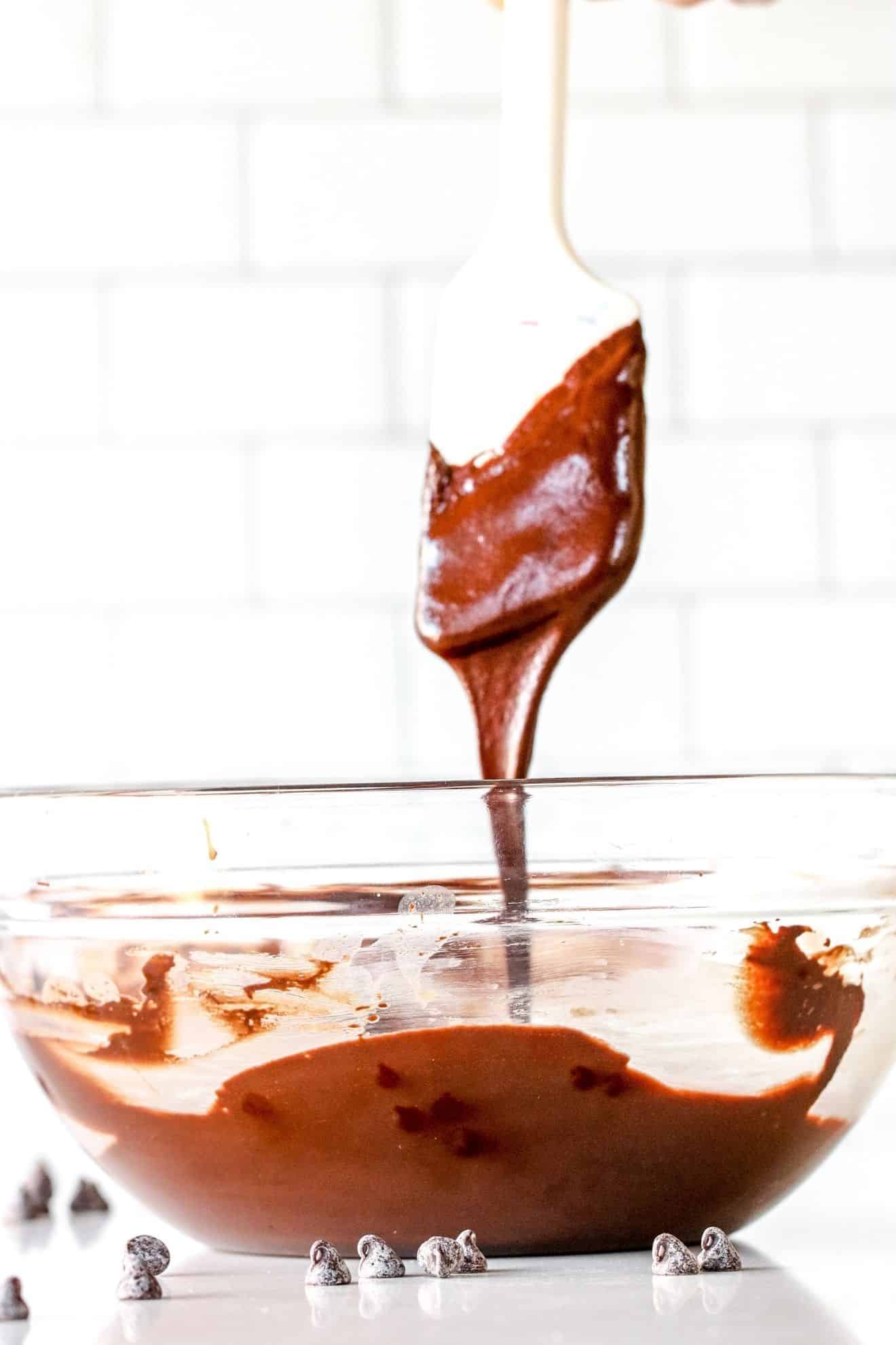 This is a side view of a glass bowl with melted chocolate in it. The bowl sits on a white counter with more chocolate chips scattered around the bowl. A white spatula is being lifted out of the bowl and drizzling the chocolate back into the bowl.