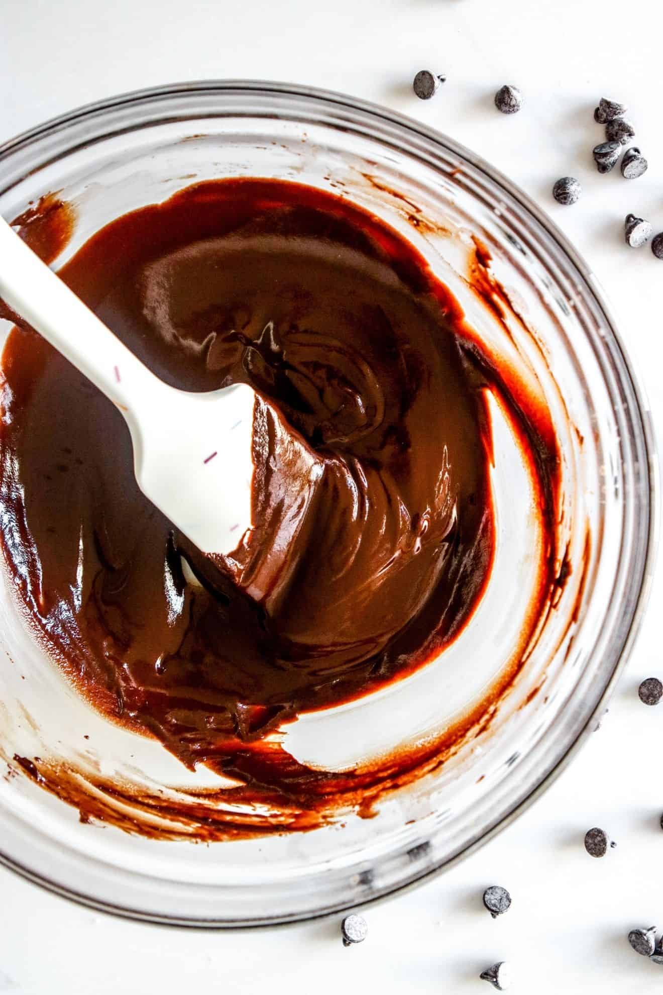 This is an overhead image of a glass bowl with melted chocolate. A spatula is in the bowl with the melted chocolate. The glass bowl sits on a white counter with more chocolate chips scattered around it.