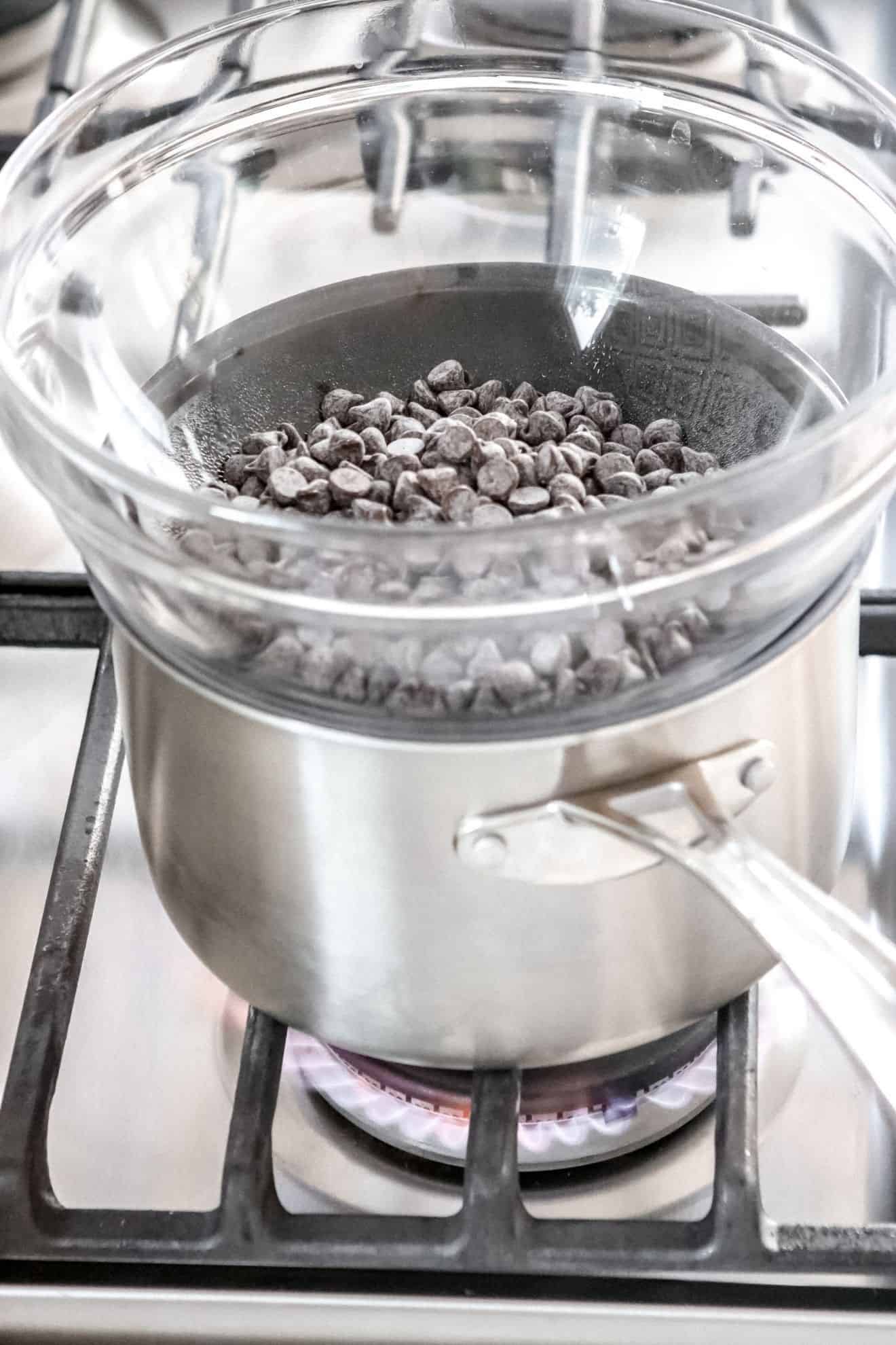 This is a side view of a stainless steel pot on a stove over a flame with a glass bowl on top. In the glass bowl are chocolate chips.