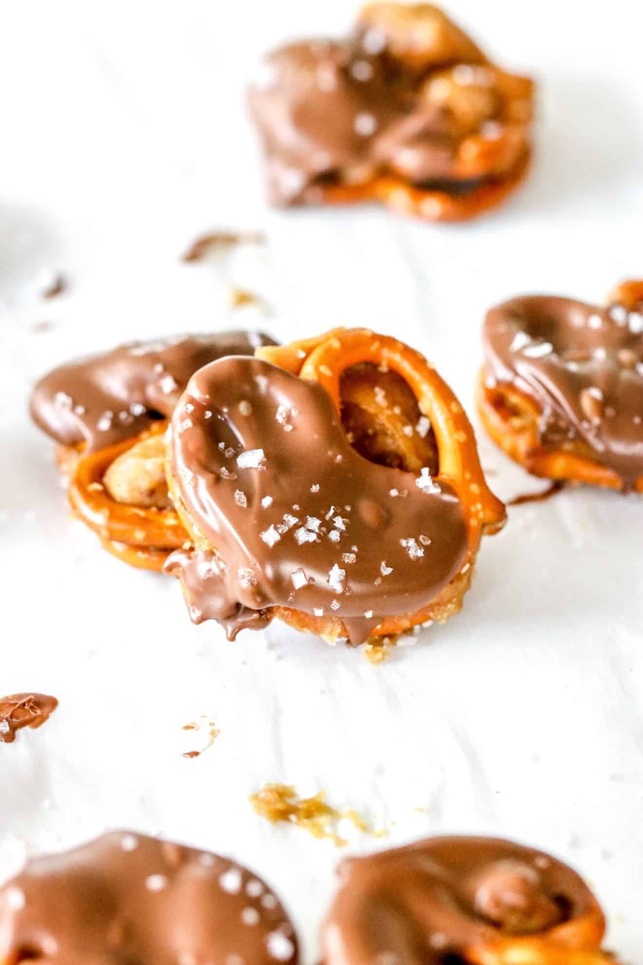 One peanut butter pretzel sandwich is dipped in chocolate and leaning against another pretzel sandwich. The pretzels wit on a white piece of parchment paper and more pretzel sandwiches are blurred in the front and behind the pretzel sandwiches.