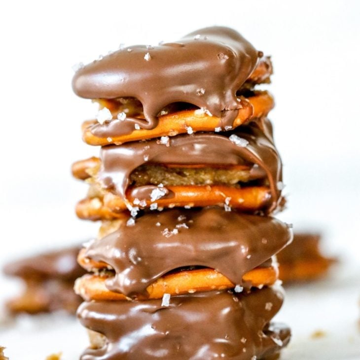 This is a stack of four pretzel sandwiches. The pretzel sandwiches have peanut butter in the middle and dipped in chocolate. They are sprinkled with salt. The stack sits on a white counter with more pretzels blurred in the background.