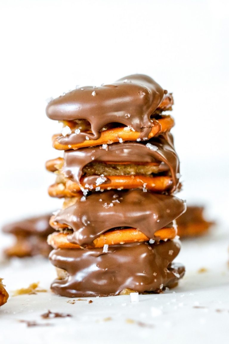 This is a stack of four pretzel sandwiches. The pretzel sandwiches have peanut butter in the middle and dipped in chocolate. They are sprinkled with salt. The stack sits on a white counter with more pretzels blurred in the background.