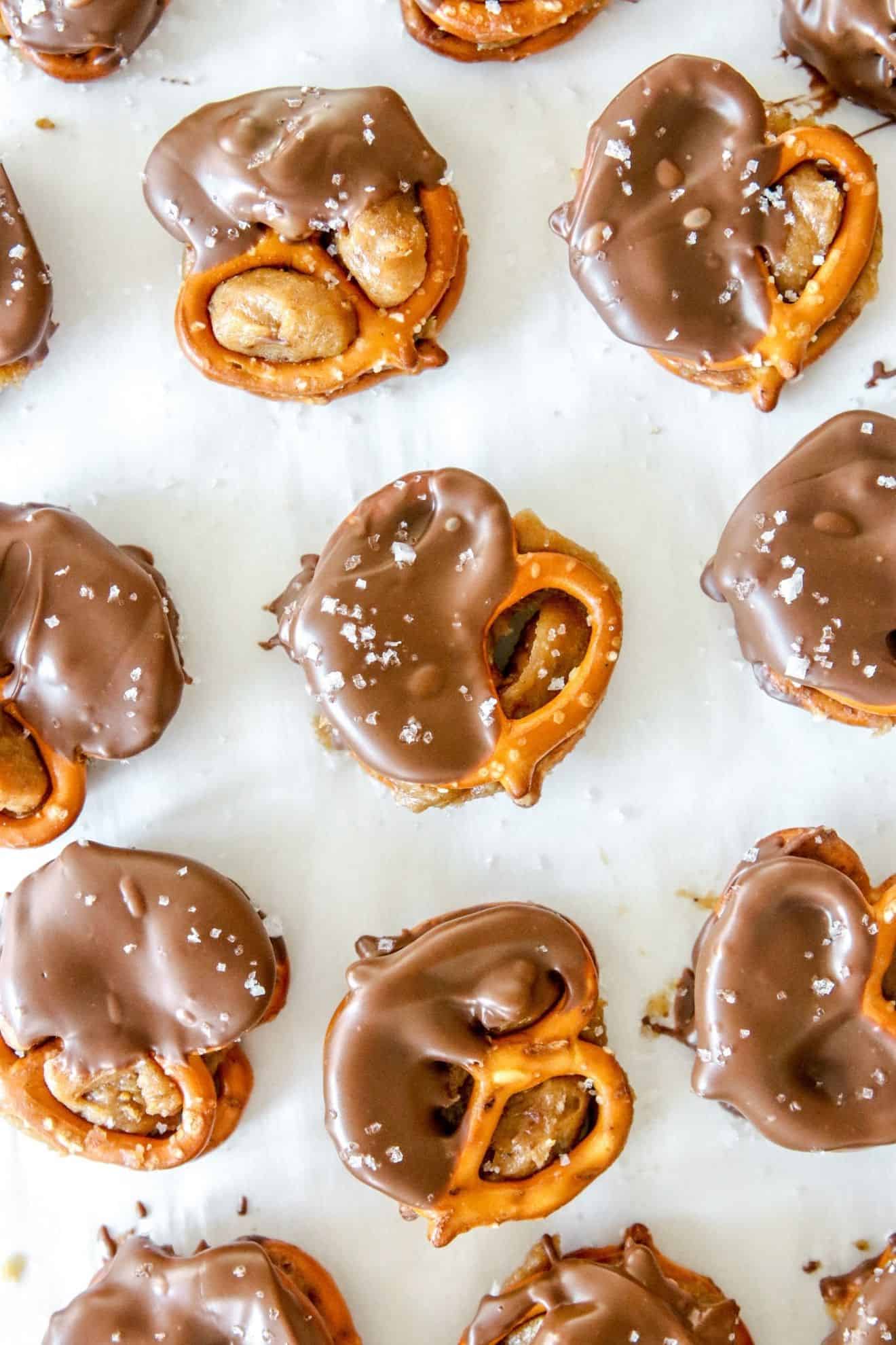 This is an overhead image of pretzels filled with peanut butter and dipped in chocolate. There is flakey salt sprinkled on top of the pretzels. The pretzels sit on a white piece of parchment paper.