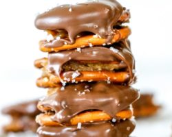 This is a stack of four pretzel sandwiches. The pretzel sandwiches have peanut butter in the middle and dipped in chocolate. They are sprinkled with salt. The stack sits on a white counter with more pretzels blurred in the background. Text overlay reads "chocolate covered peanut butter pretzels."