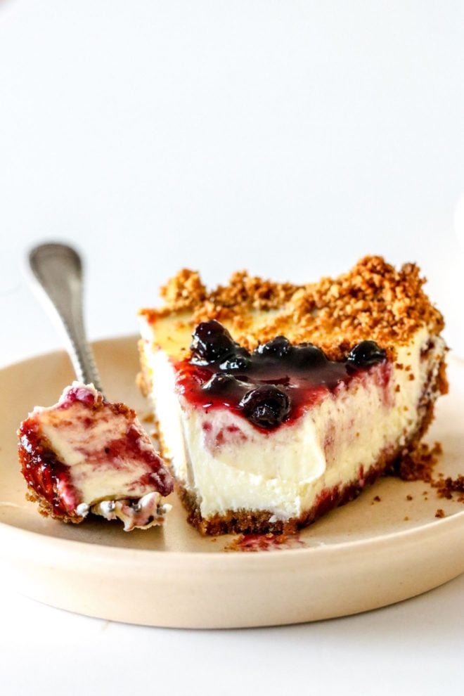 This is a side view of a slice of cream cheese pie. The slice is on a small beige plate and is topped with blueberry jam. A fork is taking off a bite of the pie and leaning on the side of the plate. The plate sits on a white counter with a white background.